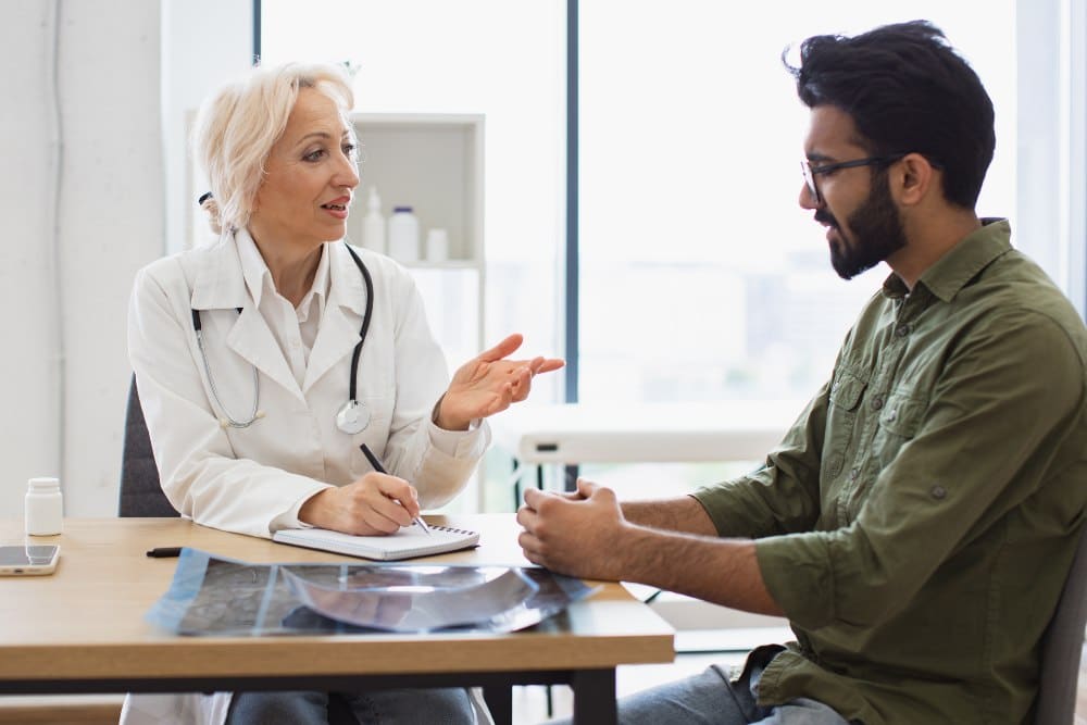 Mature elder physician giving recommendations and using notebook for writing prescription. Bearded gentleman describing symptoms of illness to senior female doctor and gesturing hands.