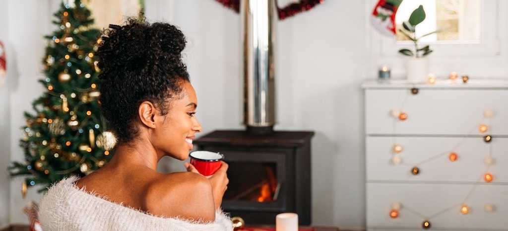 Back view of female with mug of hot tea sitting in cozy living room with Christmas decorations and fireplace