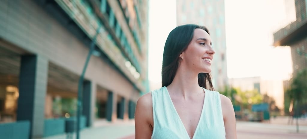 Lovely young woman with freckles and dark loose hair wearing white top and jeans is enjoying the view of modern city