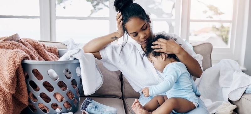 Young woman holding her crying child on the couch, laundry basket overflowing sitting next to her.