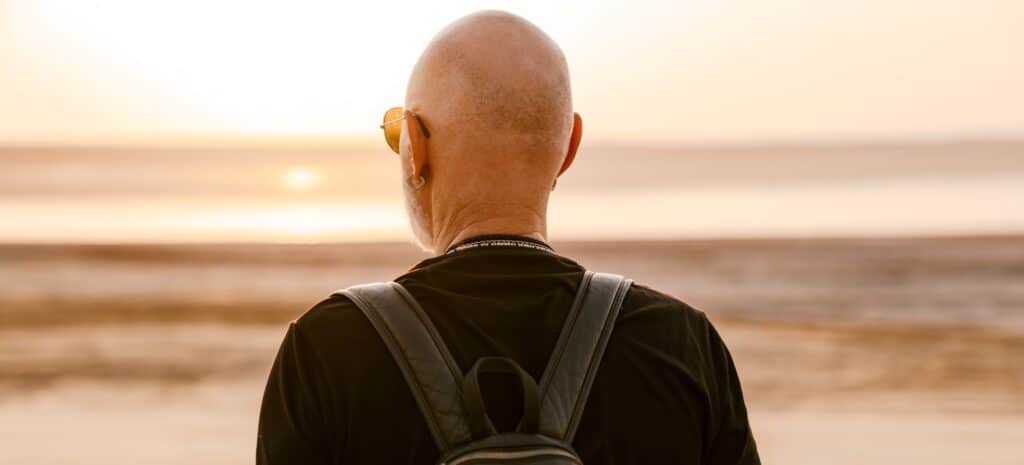 Bold senior man with backpack looking aside outdoors on summer day