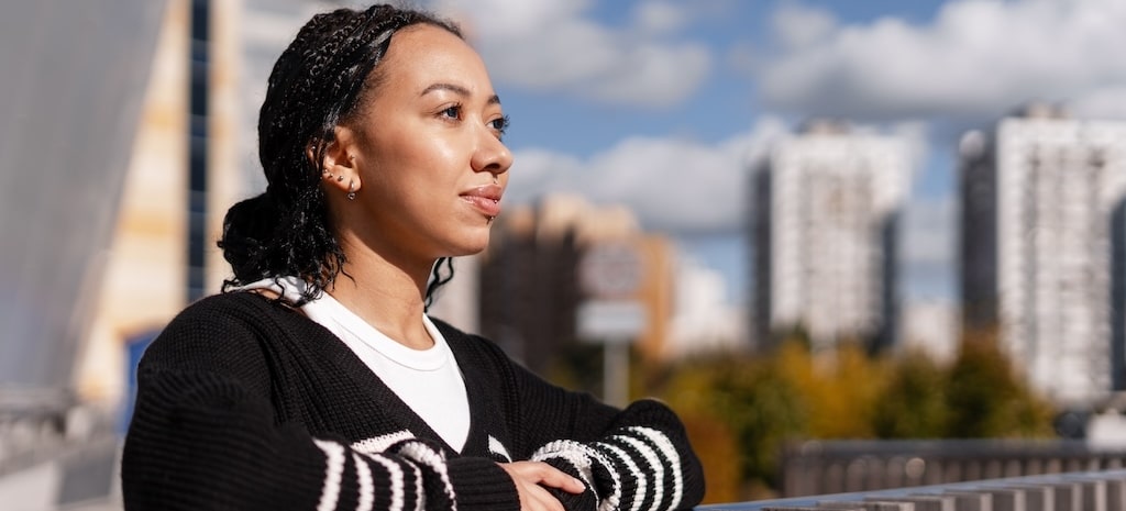 Young woman with braids looking away, thinking and contemplating while standing outdoors in an urban setting on a sunny day