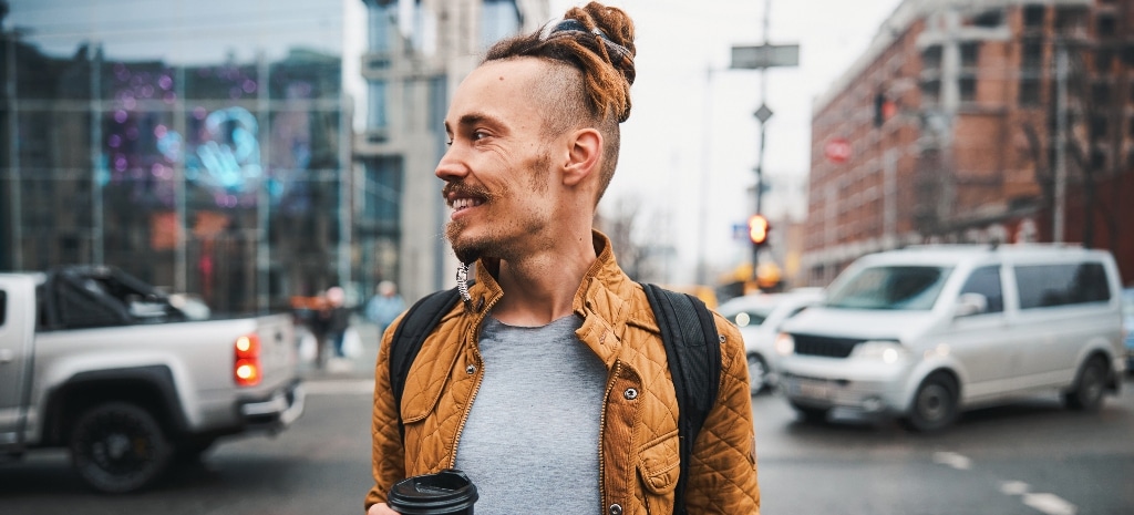 Smiling hipster with dredlocks is walking in urban roads with cup of coffee and backpack