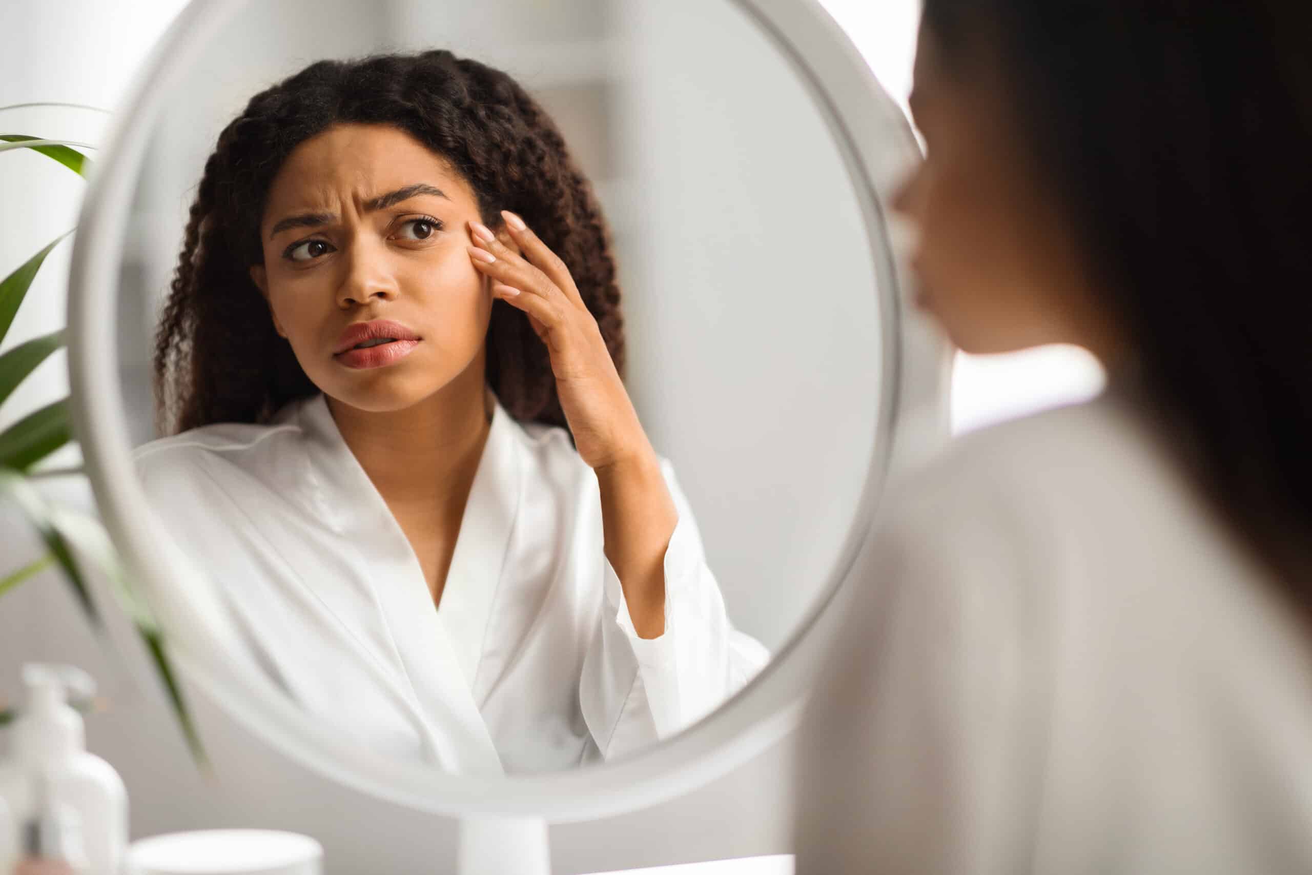 A woman examining her face in a mirror and looking concerned while wondering does alcohol make your face puffy.