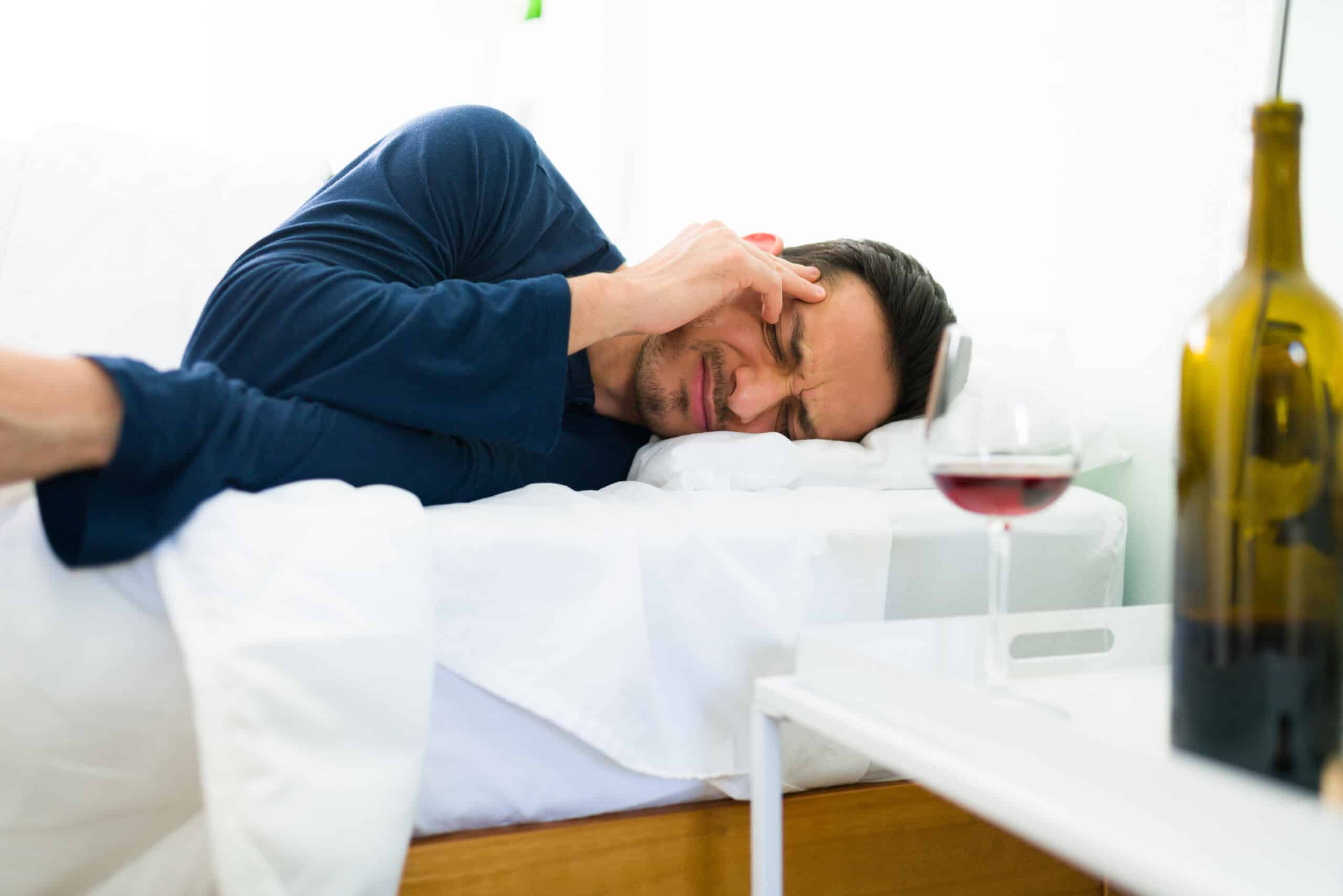 A man laying in bed hungover next to a glass and bottle of wine on his bedside table for the topic blackout drinking.