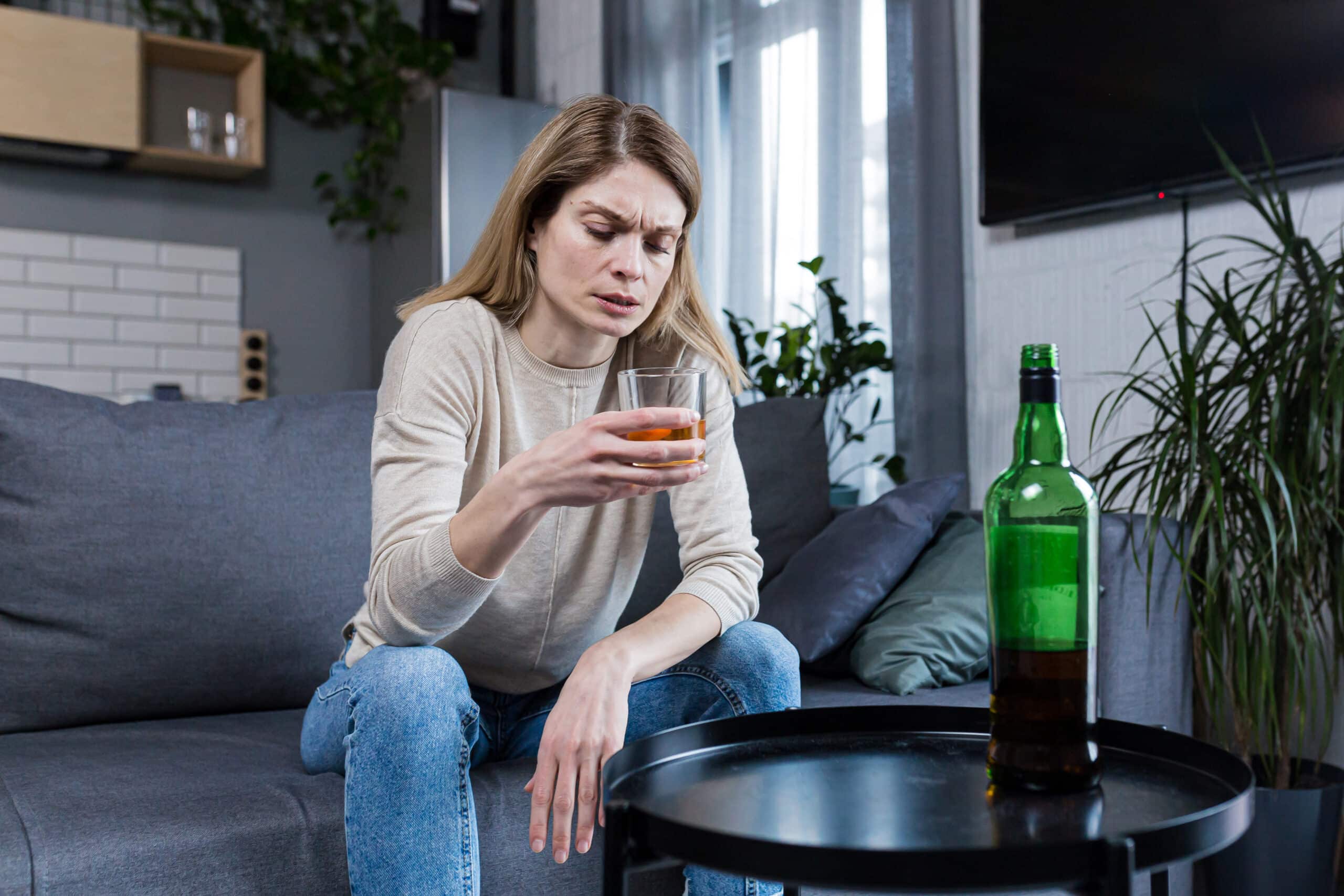 A sad woman looking sitting on her couch and looking at a glass of alcohol while asking herself is alcoholism genetic?