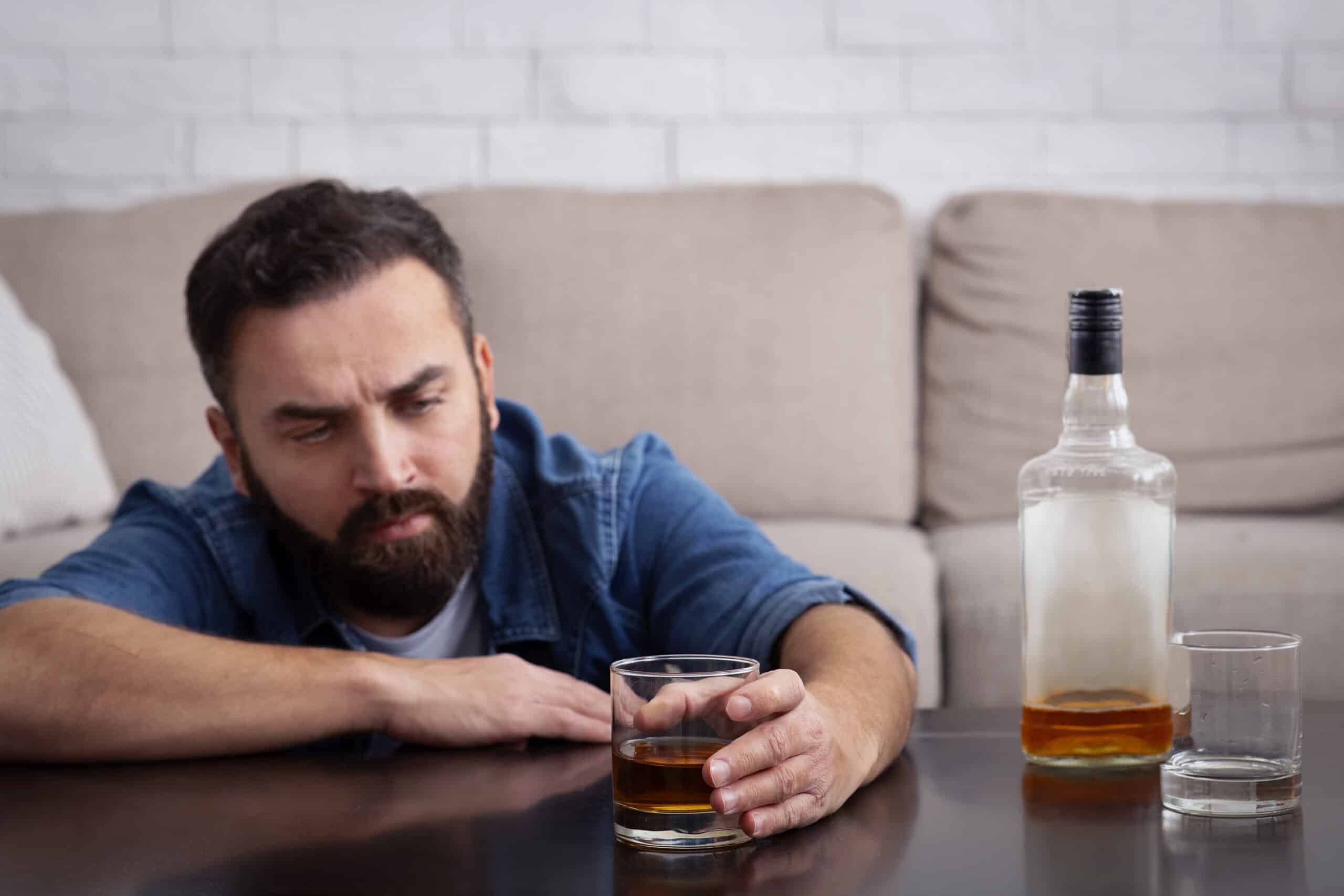 A man sitting on his floor looking sad with a glass and bottle of alcohol on the coffee table for the topic alcohol and depression.