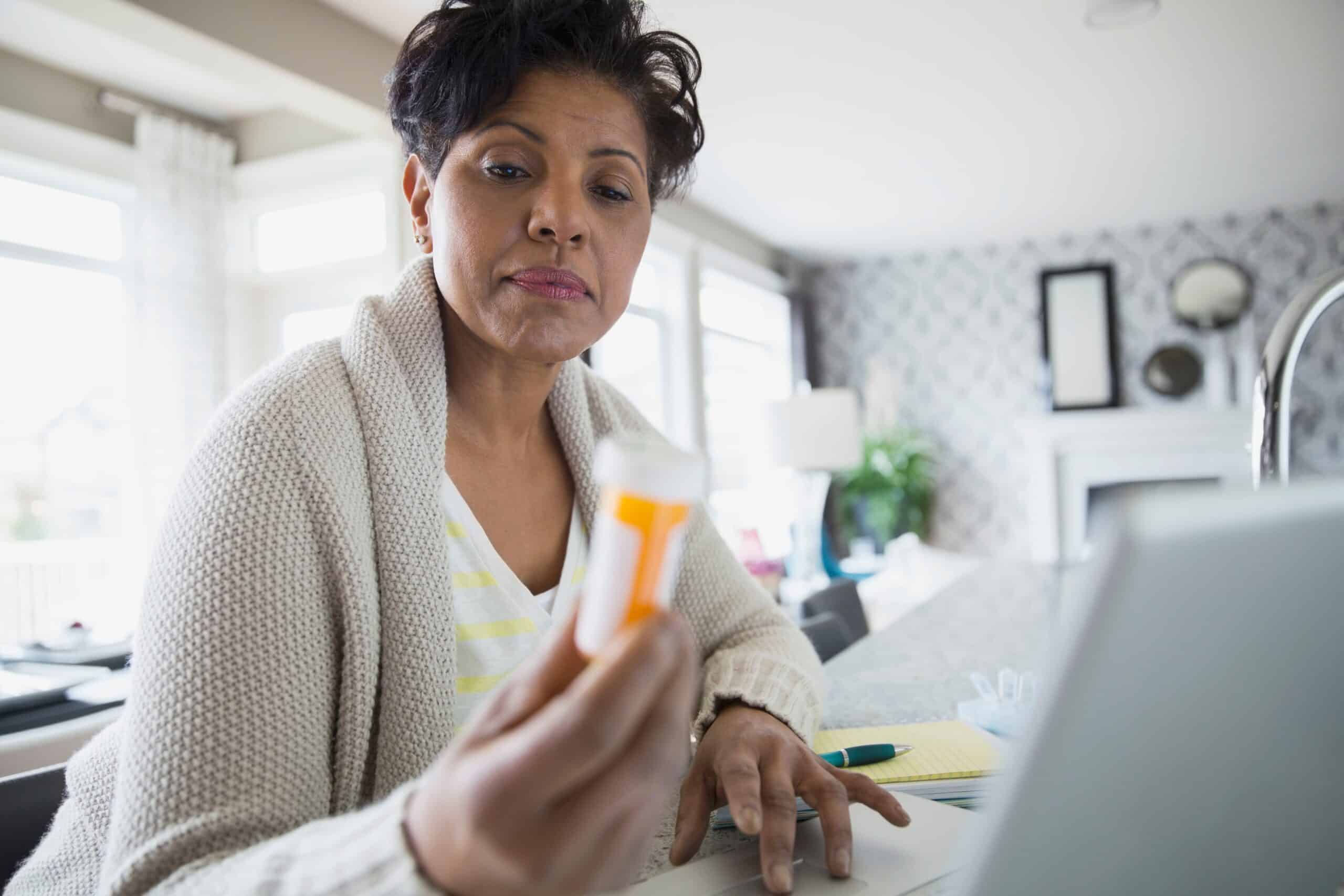A woman looks at a pill bottle while doing research on her laptop wondering about a pill to stop drinking.